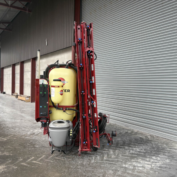 Red agricultural sprayer with a yellow tank standing inside a warehouse with corrugated metal shutters on the right side.