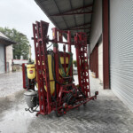 Red agricultural sprayer frame with yellow tanks and attached hoses parked beside a corrugated metal building under a roof.