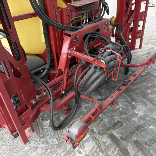 Red agricultural machinery frame with many black hydraulic hoses and wiring on a trailer; a yellow tank is visible in the background.