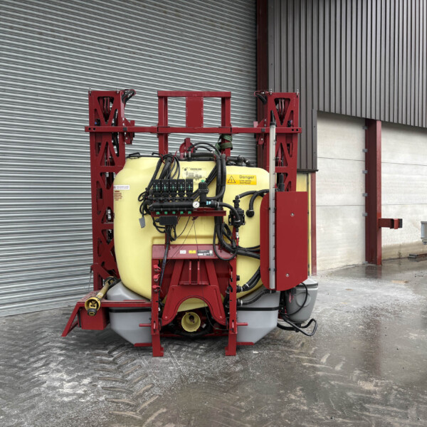 Industrial equipment with a yellow tank and red frame, hoses and gauges, mounted against a corrugated metal wall in a warehouse yard.
