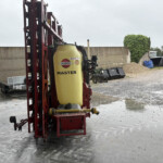Red mounted agricultural sprayer with a large yellow tank labeled HARDI MASTER, in a wet gravel yard.