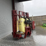 Red agricultural spray rig with a large yellow tank beside a corrugated metal wall.
