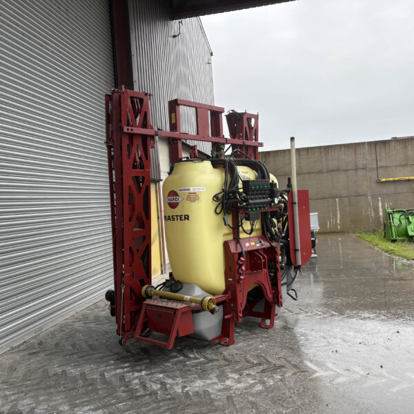 Red agricultural spray rig with a large yellow tank beside a corrugated metal wall.