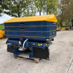 Blue industrial machine with a yellow tarp cover resting on a wooden pallet in an outdoor construction yard with trees in the background.