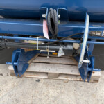 Blue agricultural equipment with coiled black hoses, metal fittings, and a measuring scale mounted on a blue frame. It sits on a wooden pallet on the ground.
