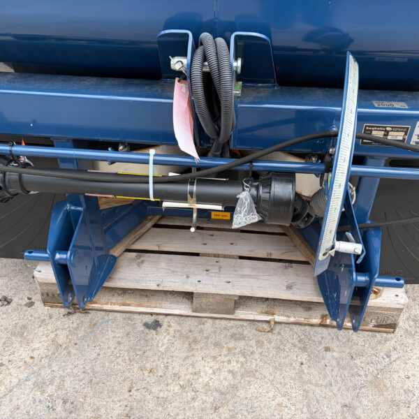 Blue agricultural equipment with coiled black hoses, metal fittings, and a measuring scale mounted on a blue frame. It sits on a wooden pallet on the ground.