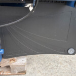 Close-up of a black equipment panel with curved grooves, a thick cable draped over it, outdoors with a blue frame and wooden pallets nearby.