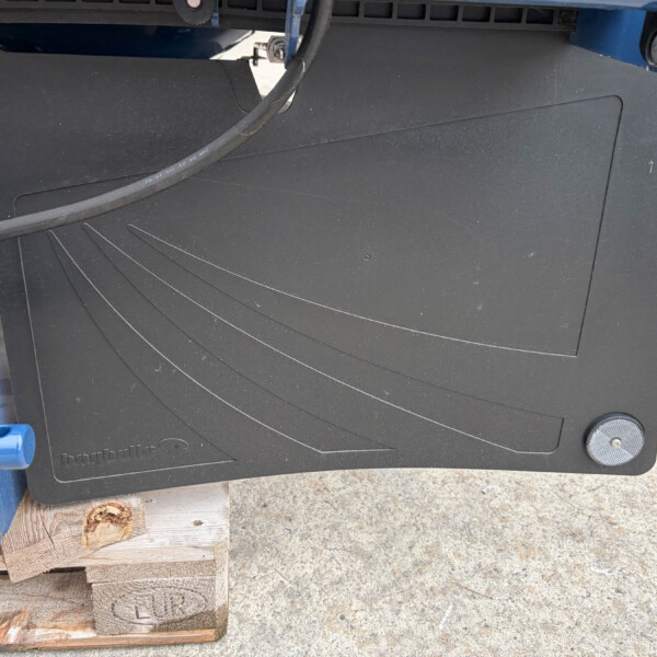 Close-up of a black equipment panel with curved grooves, a thick cable draped over it, outdoors with a blue frame and wooden pallets nearby.
