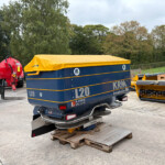 Blue KRM L20 trailer with a bright yellow cover, parked in a concrete yard among farm equipment and trees in the background.