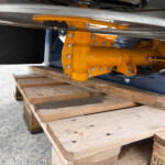 Close-up of a yellow hydraulic actuator/arm and bolt-fastened housing on industrial equipment resting on wooden pallets.
