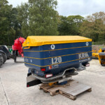 Blue transport trailer with a bright yellow cover and orange stripes, parked among construction machinery and wooden pallets in an outdoor lot.