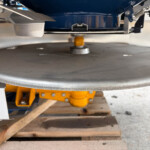 Industrial workshop close-up: yellow support pillar under a gray rotating turntable with metal arms and a blue machine above. Resting on wooden pallets in the background.