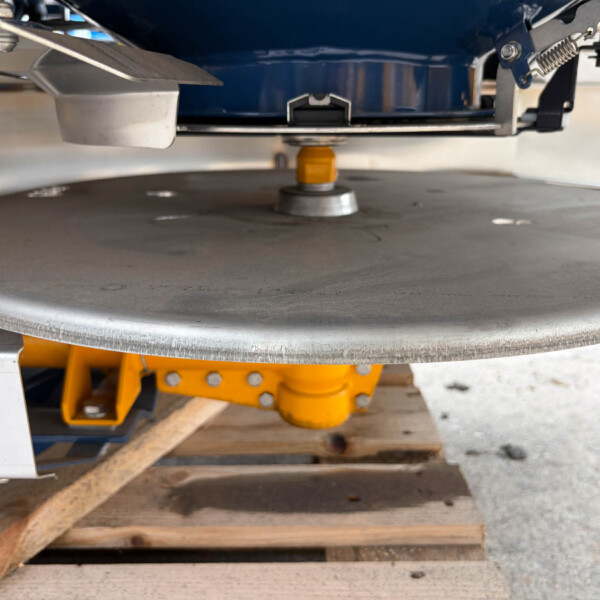 Industrial workshop close-up: yellow support pillar under a gray rotating turntable with metal arms and a blue machine above. Resting on wooden pallets in the background.
