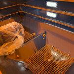 Interior of a dark blue industrial cabinet or compartment with orange safety trim, plastic-wrapped component on a metal grate floor, and built-in lights along the wall