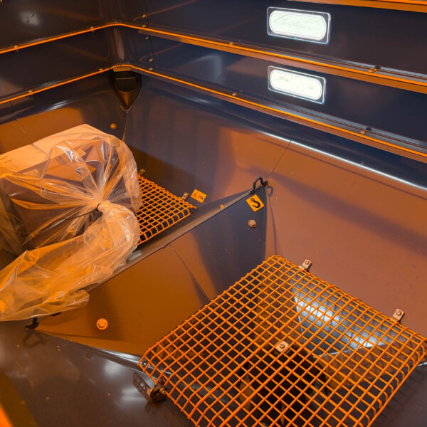 Interior of a dark blue industrial cabinet or compartment with orange safety trim, plastic-wrapped component on a metal grate floor, and built-in lights along the wall