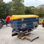 Blue trailer with a yellow-covered load labeled 'L20' in a farm equipment yard, surrounded by tractors and other machinery, with trees in the background.