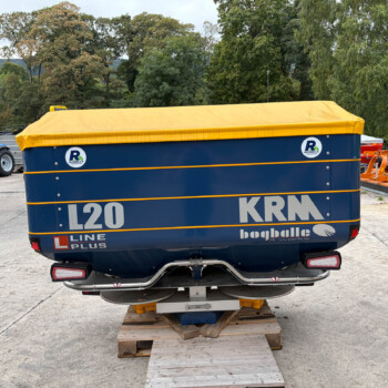 Large blue trailer/vehicle with a yellow cover on a wooden pallet, outdoors in an industrial yard with tractors and equipment nearby and trees in the background