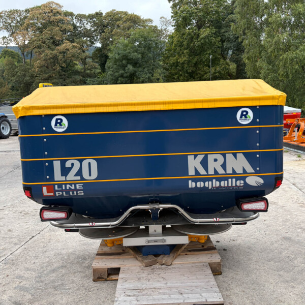 Large blue trailer/vehicle with a yellow cover on a wooden pallet, outdoors in an industrial yard with tractors and equipment nearby and trees in the background
