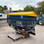 Blue trailer with a yellow tarp cover in an industrial yard; KRM branding visible, with a white van and farming equipment nearby.