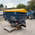 Blue trailer with a yellow tarp cover parked on wooden pallets in an outdoor yard; a white van and farm equipment are in the background.