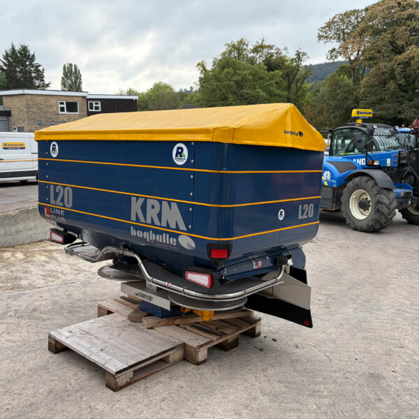 Blue trailer with a yellow tarp cover parked on wooden pallets in an outdoor yard; a white van and farm equipment are in the background.