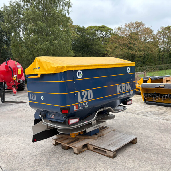 Blue KRM L20 trailer with a bright yellow cover, parked in a concrete yard among farm equipment and trees in the background.