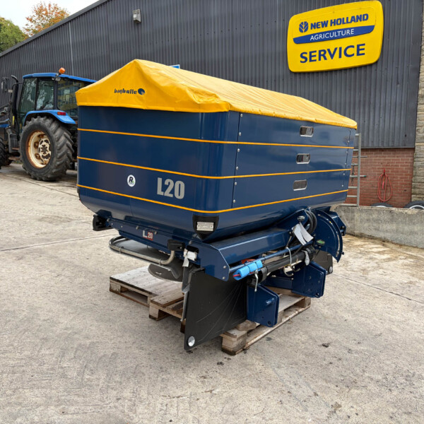 Blue L20 farming trailer with a yellow tarp cover parked beside a tractor near a New Holland Agriculture Service sign.