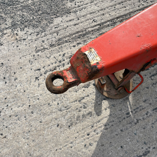 Close-up of a red metal hydraulic component resting on a gravelly concrete surface, showing a circular hole and worn, chipped paint.