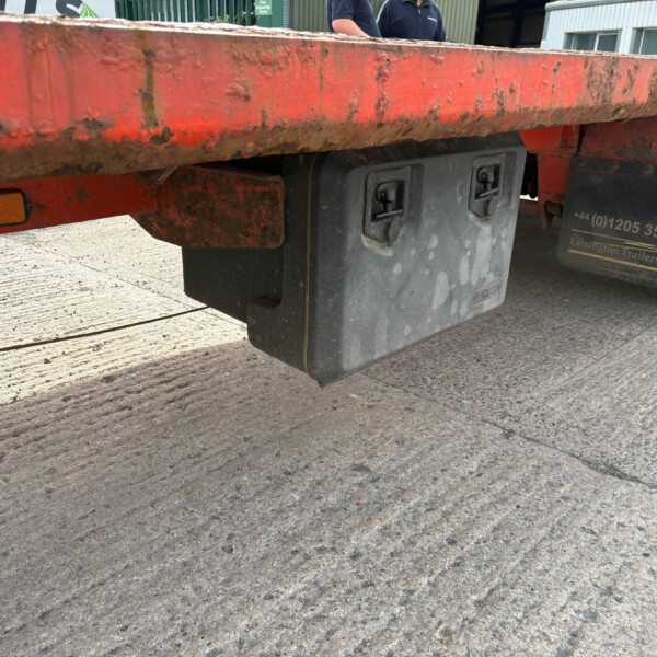 Close-up of the underside of a red, dirty trailer showing a black toolbox and mud flaps under the frame.