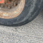 Close-up of a rusted wheel hub and worn tire on rough pavement, showing corrosion and bolts at the wheel's edge