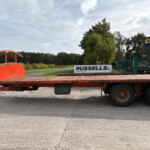 Red flatbed trailer in the foreground with a New Holland Agriculture building and trees in the background.