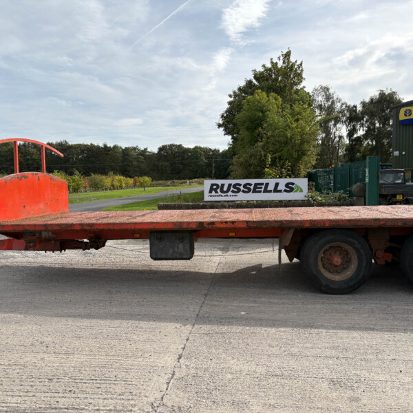 Red flatbed trailer in the foreground with a New Holland Agriculture building and trees in the background.