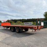 Red flatbed trailer in an industrial lot with a blue car parked nearby and green fence in the background, signage reading 'RUSSELLS' visible.