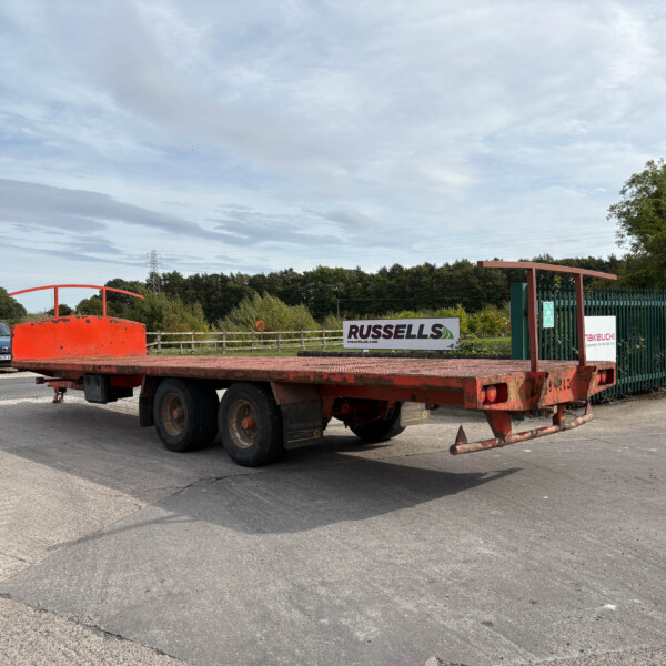 Red flatbed trailer in an industrial lot with a blue car parked nearby and green fence in the background, signage reading 'RUSSELLS' visible.