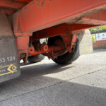 Red trailer underside with wheels and rusted metal frame, viewed from ground level. Mud flaps show company markings and safety icons.