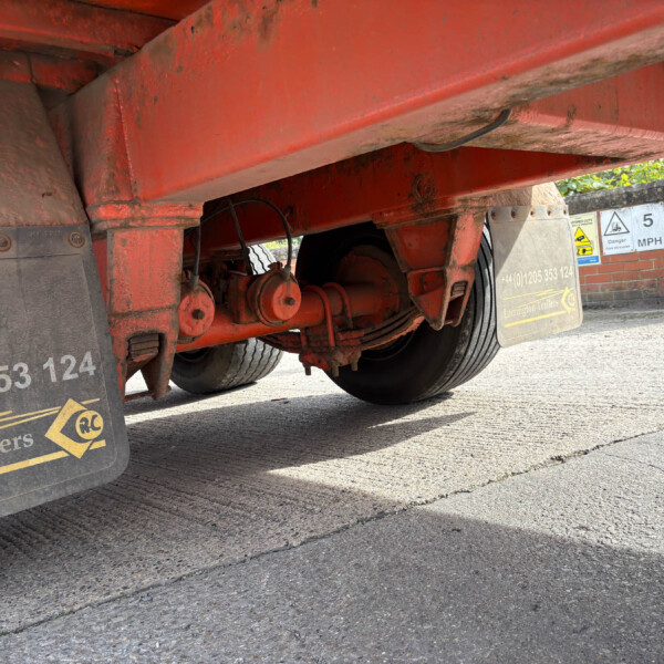 Red trailer underside with wheels and rusted metal frame, viewed from ground level. Mud flaps show company markings and safety icons.