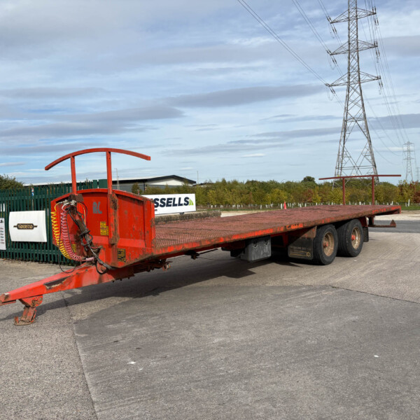 Red flatbed trailer with equipment in an industrial lot, power lines and pylons in the background under a cloudy sky