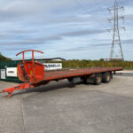 Red flatbed trailer with equipment sits in an industrial yard, with tall power pylons and transmission lines in the distance under a cloudy sky.