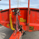 Rear of a red industrial trailer with coiled yellow and pink hoses and warning labels, outdoors near a green fence.