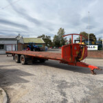 Red heavy farm equipment on a flatbed trailer in an outdoor yard, with a blue tractor and New Holland flags nearby under a cloudy sky