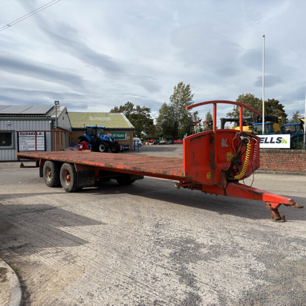 Red heavy farm equipment on a flatbed trailer in an outdoor yard, with a blue tractor and New Holland flags nearby under a cloudy sky