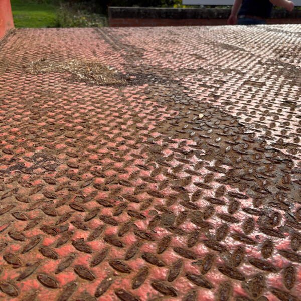 Close-up of a rusted red diamond-plate metal surface with mud, outdoors, a person partially visible in the background.