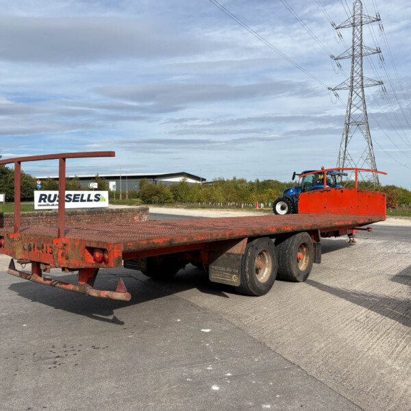Red flatbed trailer in an industrial yard with a blue tractor nearby and power lines overhead.