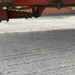 Underside of a red trailer showing its frame, suspension, and hydraulic components over a concrete surface near a brick wall.