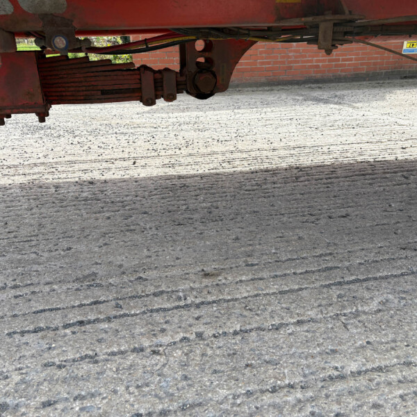 Underside of a red trailer showing its frame, suspension, and hydraulic components over a concrete surface near a brick wall.