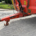 Close-up of red industrial machinery with a yellow spiral hose and warning labels on a rough pavement surface.