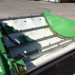 Close-up of a green farming header with sharp metal teeth along the edges, resting on a concrete surface.