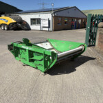 Green agricultural machine in the foreground at an industrial yard, with a yellow New Holland machine and a Russell's building in the background on a sunny day.