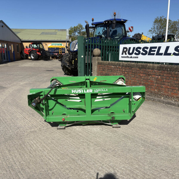Green Hustler Unrolla LX105 mower in the foreground at a farm equipment yard, with a Russell's sign on a green fence to the right.