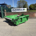 Blue tractor and a green farming implement parked behind a brick wall with a Russell’s sign in a yard under a clear blue sky.
